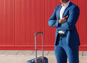 Crop unrecognizable black businessman in classy blue suit standing with arms crossed near suitcase against red wall