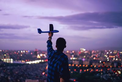 Rear view of man holding model airplane against illuminated cityscape at dusk