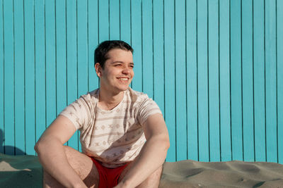 Young man wearing swimsuit pants smiling in sandbeach looking at sun