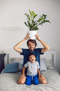 Bearded father and son in t-shirts are sitting on a bed. man is holding pot with flower on his head