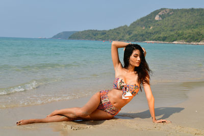 Young woman wearing bikini sitting at beach against clear sky