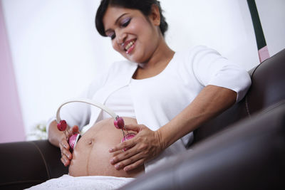 Pregnant woman holding headphones on belly while sitting on sofa at home