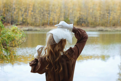 Young blonde woman in vintage clothes standing near lake in autumn park.