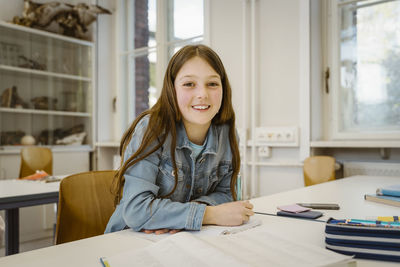 Portrait of smiling schoolgirl with long hair sitting at desk in classroom