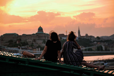 Rear view of female friends sitting on liberty bridge in city against orange sky