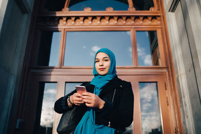 Low angle portrait of confident young muslim woman standing with smart phone against entrance door