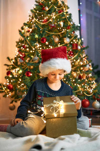 A charming boy in a santa claus hat sits next to the christmas tree and opens a box with a gift. 