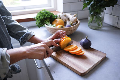 Midsection of woman slicing persimmon on cutting board at kitchen counter