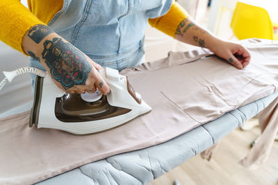 Cropped unrecognizable female in overall ironing clothes on board during housework routine in light room at home