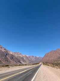 Road leading towards mountains against clear blue sky