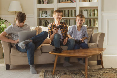 Three boys, brothers, sit on the couch at home in the sunlight and play computer and video games.