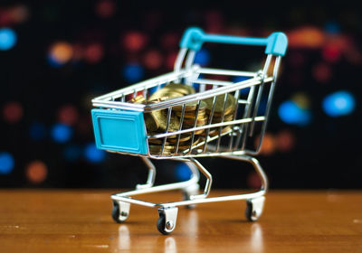 Close-up of miniature shopping cart with coins on table against lights