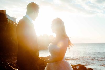 Couple holding hands while standing by sea against sky during sunny day