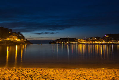 Illuminated buildings by sea against sky at night