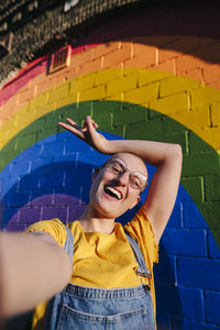 Cheerful transgender person gesturing peace sign and taking selfie in front of rainbow wall