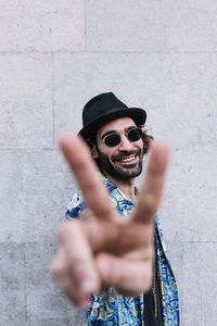 Smiling young man showing peace sign while standing in front of wall