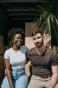 Portrait of happy multiracial couple sitting with arms around in van trunk