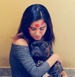 Woman with lipstick kiss on face holding dog against wall at home