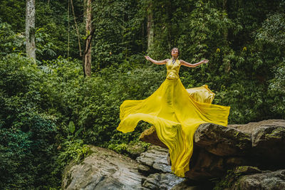 Full length of young woman in long yellow dress standing on cliff in forest
