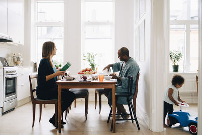 Man and woman having breakfast together at dining table while daughter playing in living room