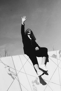 Low angle view of young woman shielding while sitting on retaining wall against sky during sunny day