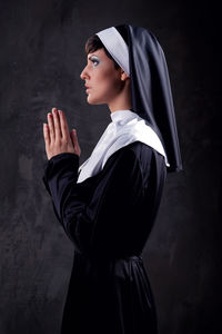 Side view of nun praying while standing against black background