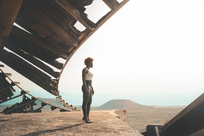 Full body side view of slim thoughtful african american female in trendy outfit and high heels standing near shabby wooden construction ind desert area on summer day