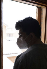 Young man with mask in quarantine looking at the snowfall through the window of his house