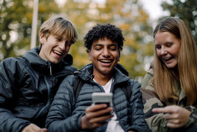 Happy teenage boy showing smart phone to male and female friends in city
