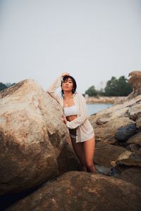 Portrait of young woman wearing bikini standing by rocks near sea against clear sky