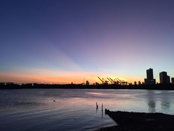 Silhouette buildings by sea against sky at sunset