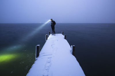Hiker with headlamp standing on snow covered jetty at dusk, walchensee, bavaria, germany