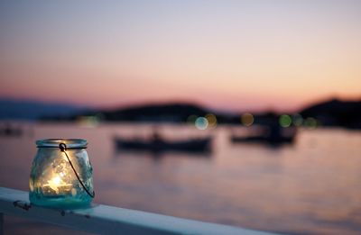 Lit candle in glass jar on railing by river against clear sky during sunset