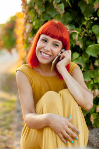 Smiling woman talking on phone while sitting against plant