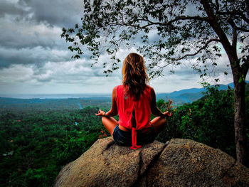 Rear view of woman meditating on rock by forest against cloudy sky