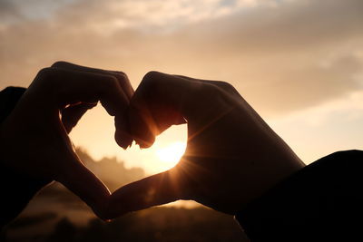 Hand holding heart shape against sky during sunset