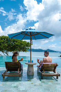 Rear view of woman sitting on lounge chair at beach against sky