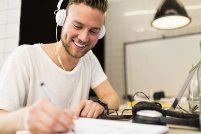 Happy young businessman wearing headphones while writing in book at creative office