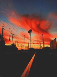 Silhouette electricity pylon by road against dramatic sky during sunset