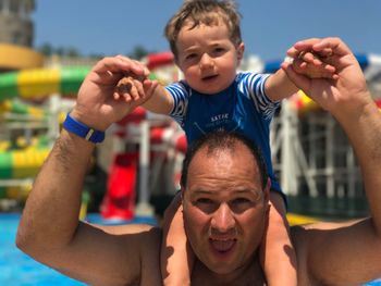 Portrait of shirtless father carrying son on shoulders at water park during sunny day