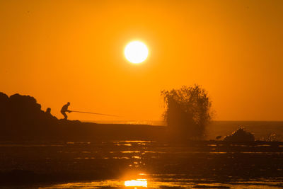 Silhouette man fishing at beach against orange sky