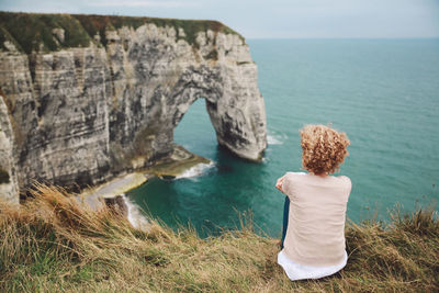 Rear view of woman sitting on field by sea against sky