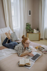 Smiling woman with coffee cup using laptop lying on bed at home