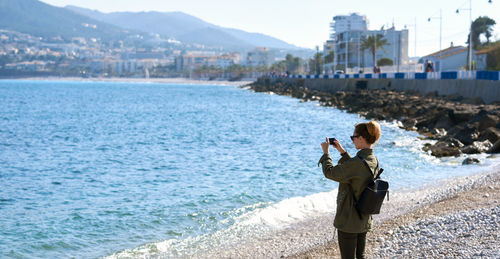 Woman photographing through mobile phone while standing at beach