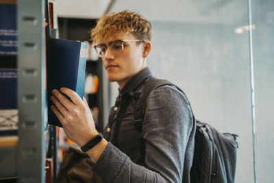 Side view of young man wearing eyeglasses searching book in library at university
