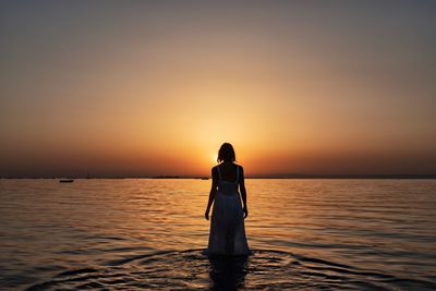 Rear view of woman standing in sea against sky during sunset