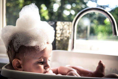 Close-up of cute baby boy with soap sud on head in kitchen sink