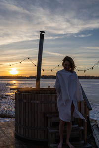 Rear view of woman standing at beach against sky during sunset