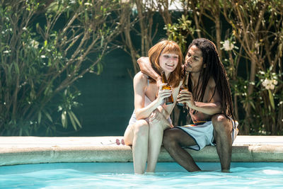 Couple toasting beer bottles while sitting at poolside