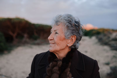 Happy elderly female tourist with gray hair in warm casual outfit smiling and looking away while relaxing on sandy beach against cloudy evening sky